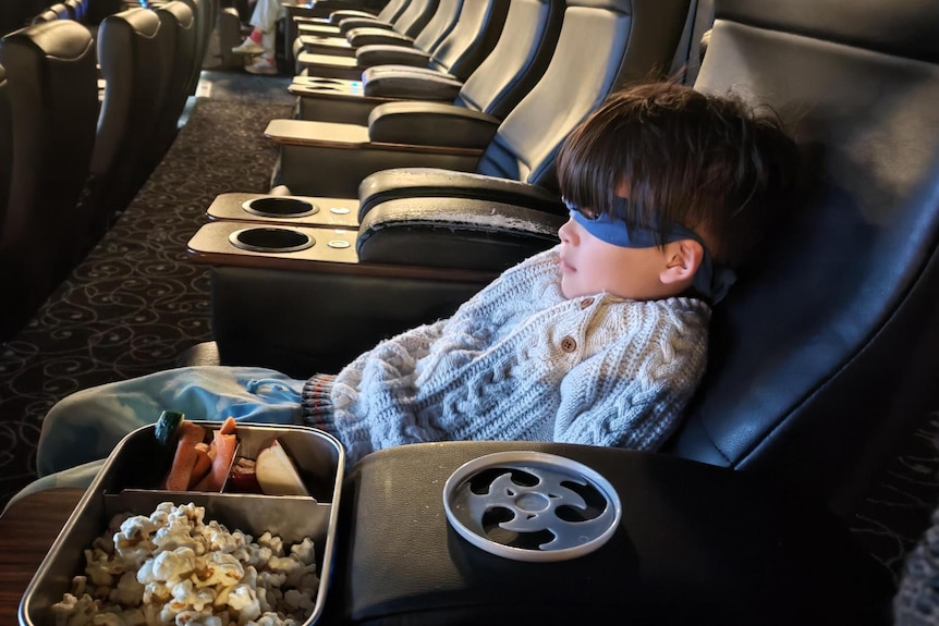 Young boy in turtle mask leans back in a cinema chair, with popcorn and ninja star seated beside him 
