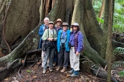 Five women in long pants and shirts and hats, stand in a rainforest with binoculars.
