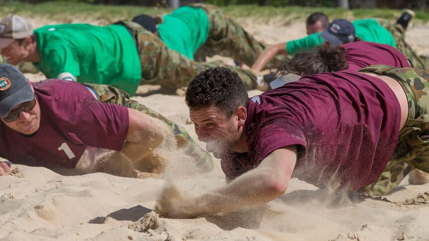 A man in a coloured t-shirt army crawls through sand as another man looks on.