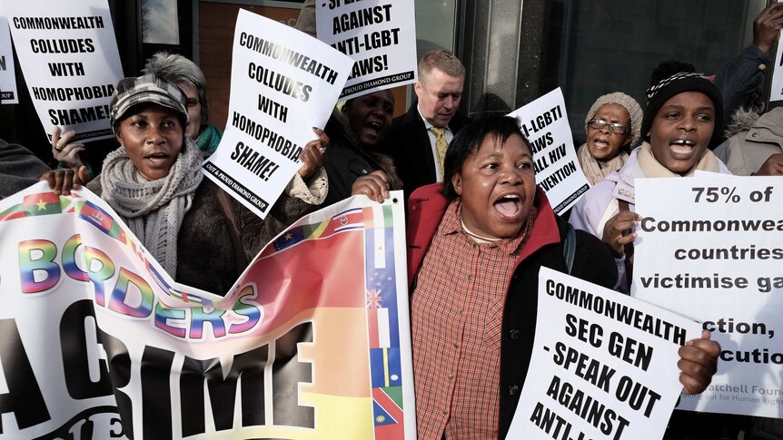 A group of people protest outside of glass doors hold white placards that say 'Commonwealth colludes with homophobia'