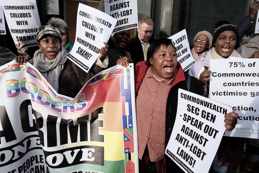 A group of people protest outside of glass doors hold white placards that say 'Commonwealth colludes with homophobia'