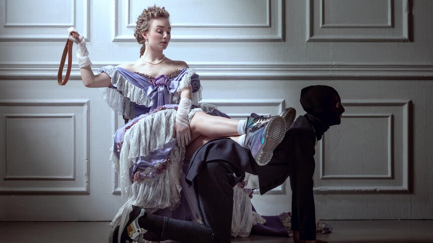 A woman in a period costume holding a riding crop sits with her feet up, using a hooded person on all fours as a foot stool