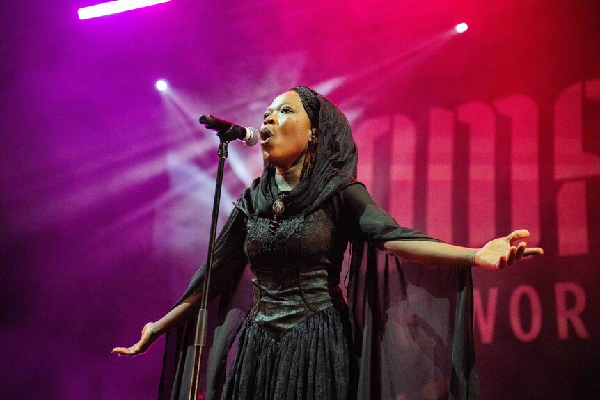 A woman in black dress and headscarf sings on stage with her arms outstretched