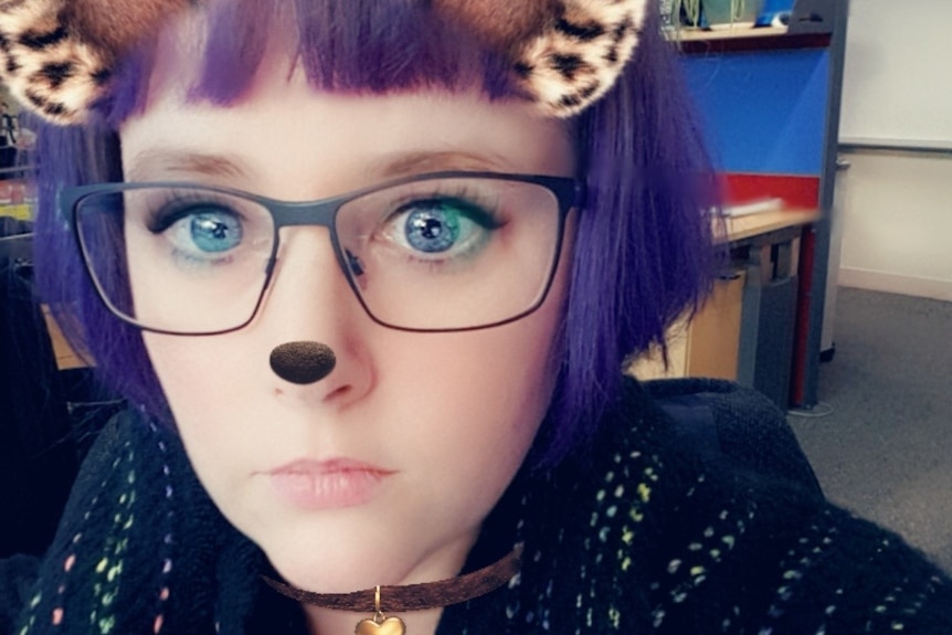 A selfie showing a woman with purple hair with cartoon eyes, ears and a black dot on her nose