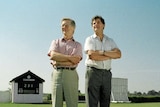 Two men stand on grass with arms folded, with a cricket scoreboard and sightscreen in background.