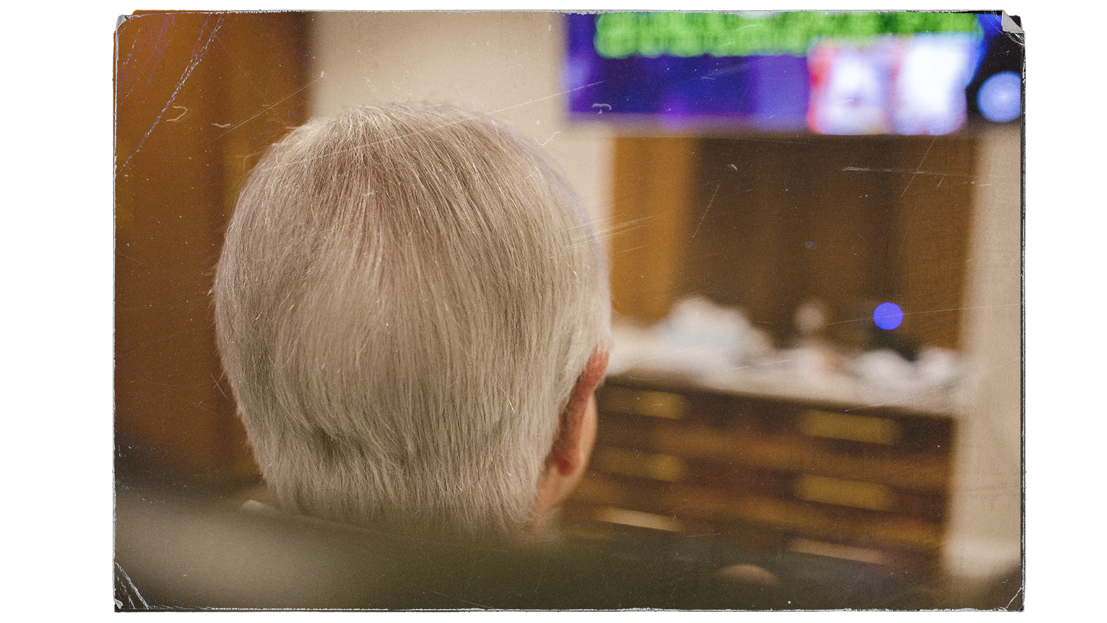 The back of a man's head with grey hair with a tv going in the background