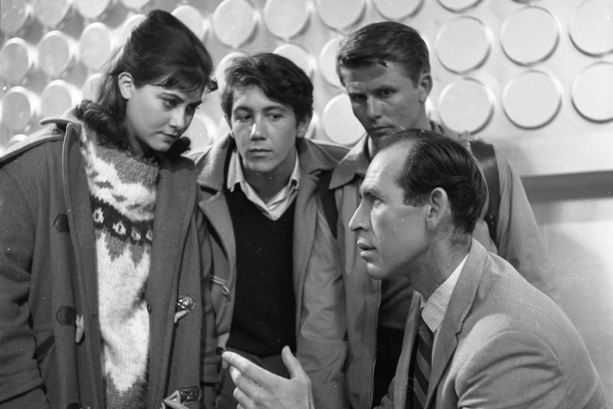 A man in a suit talking with three children in a room with futuristic interior. The picture is in black and white.