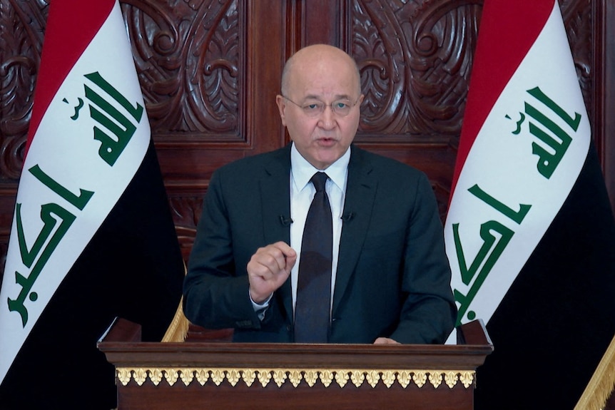 A man in a suit stands at a lectern, flanked by two Iraqi flags. He pinches to gesture as he speaks