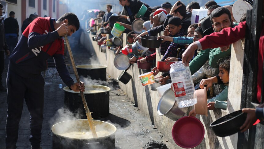 A man stirs a large pot of food while people on the other side of a barrier hold out receptacles. 