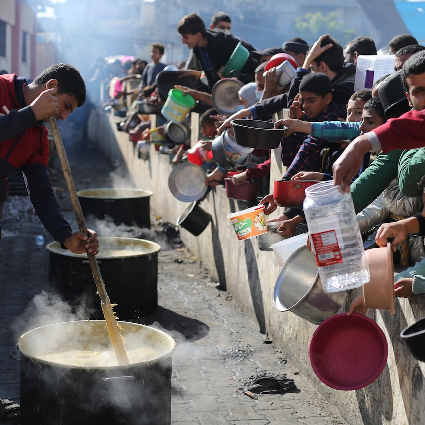 A man stirs a large pot of food while people on the other side of a barrier hold out receptacles. 
