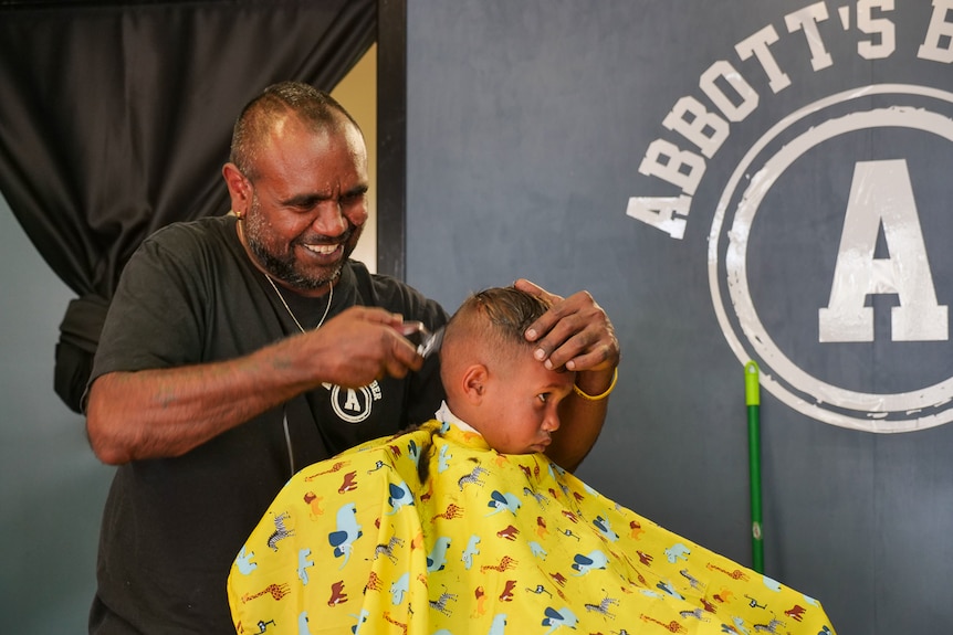 A smiling Indigenous man gives a boy a haircut in a barber shop.