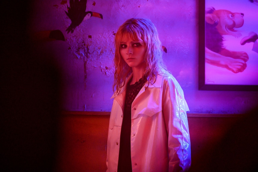Bathed in a purple light, a woman in white trenchcoat and wet blonde hair