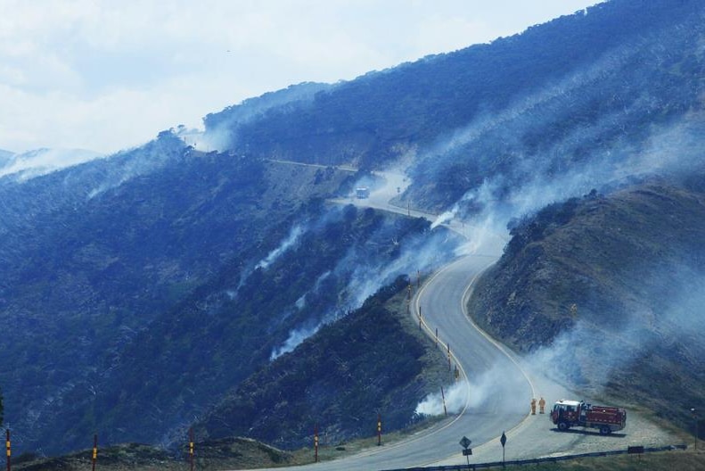 Smoke comes from a steep mountainside, and firefighters are stationed on a road at the top of a mountain with a truck