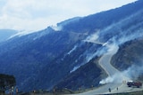 Smoke comes from a steep mountainside, and firefighters are stationed on a road at the top of a mountain with a truck