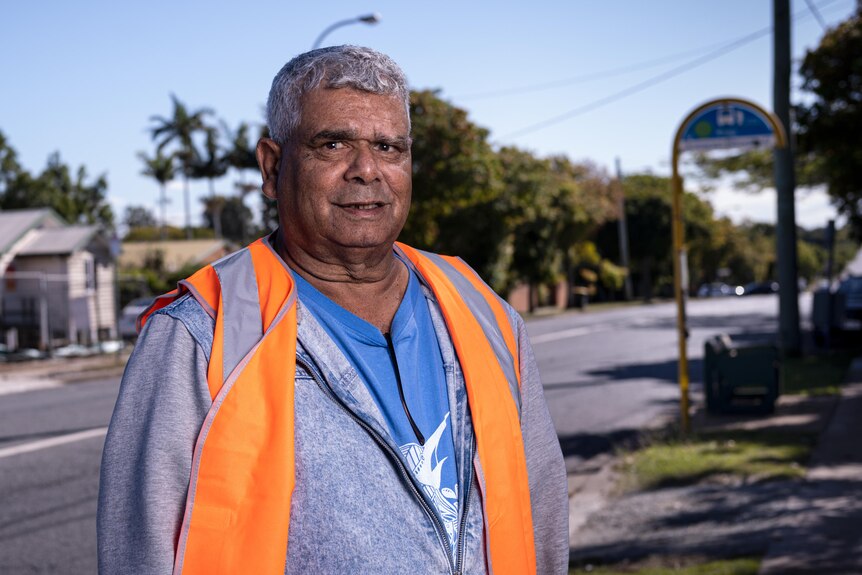 A smiling Indigenous man in a a high-vis vest stands on a sunny street.