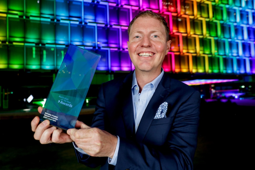 A man in a suit holds up a glass award in front of a building lit up rainbow.