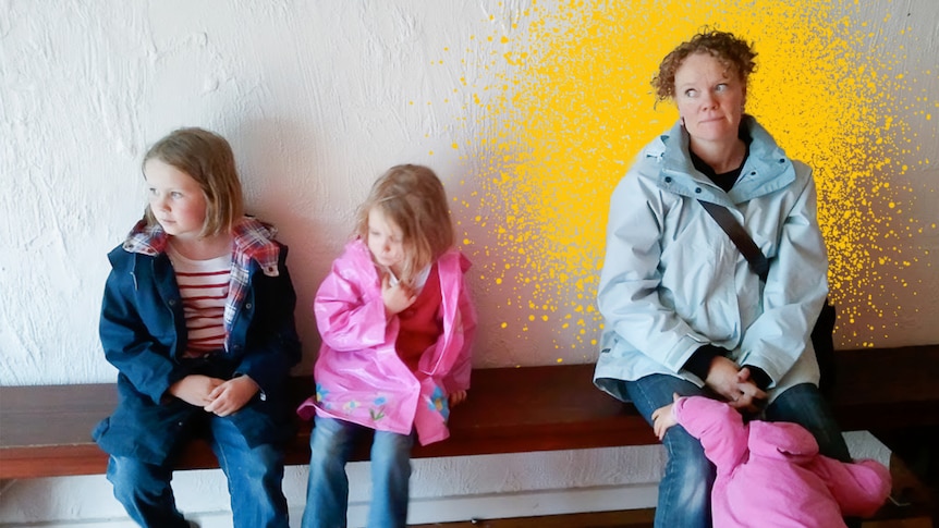 Mandy McCracken sits on a bench indoors with her two daughters. They wear rain clothes and look to the left.