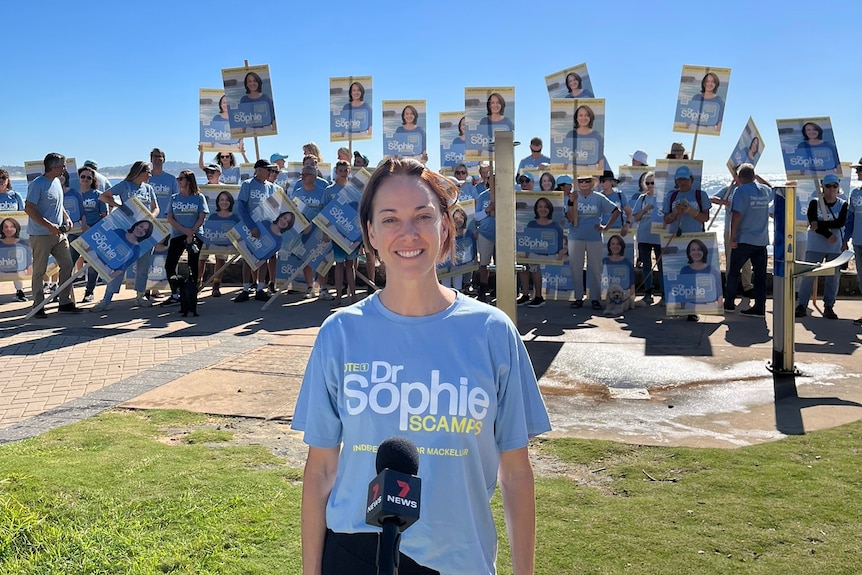 A woman stands smiling outdoors with a crowd of supporters behind her holding signs with her face.