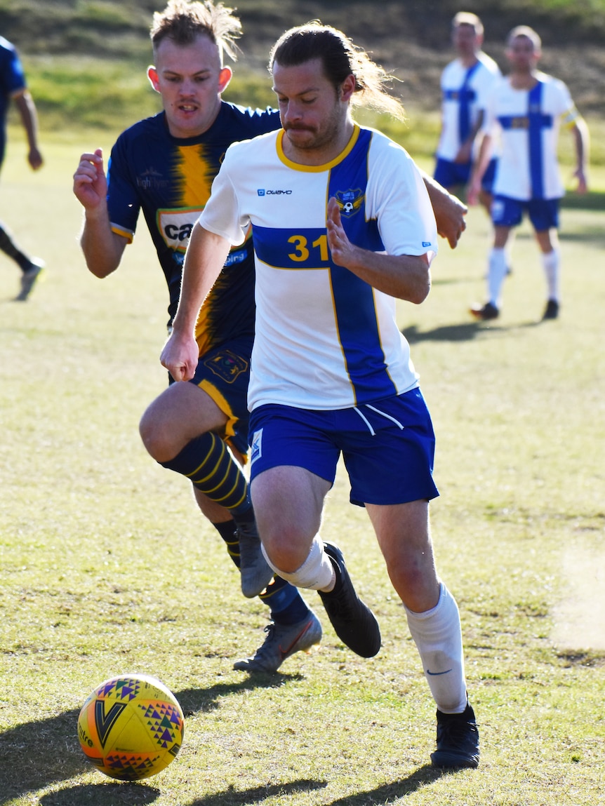 A footballer looks down as he runs with the ball, while an opponent runs close behind him.