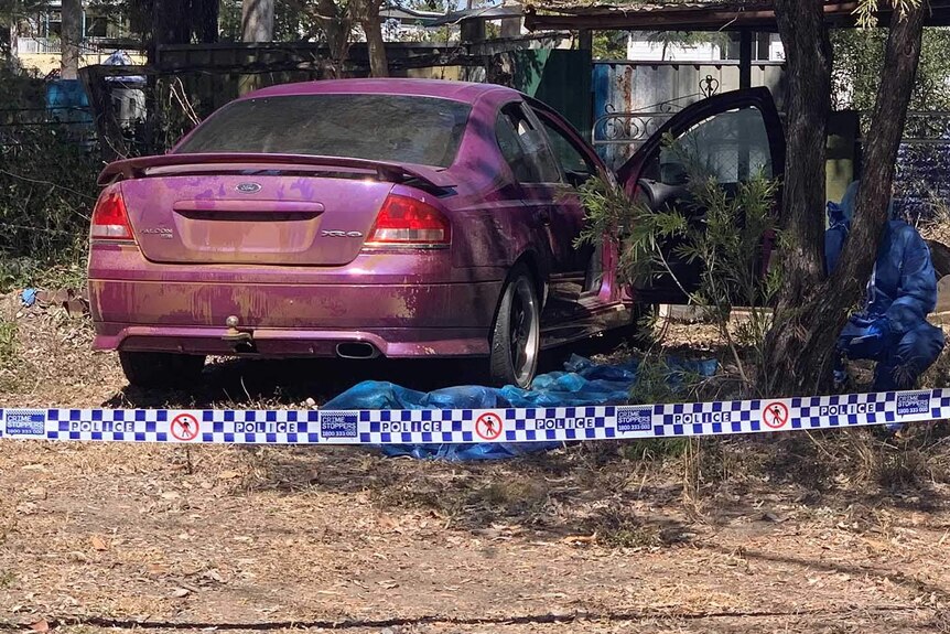 A purple sedan abandoned in bush near a wire fence being photographed by police