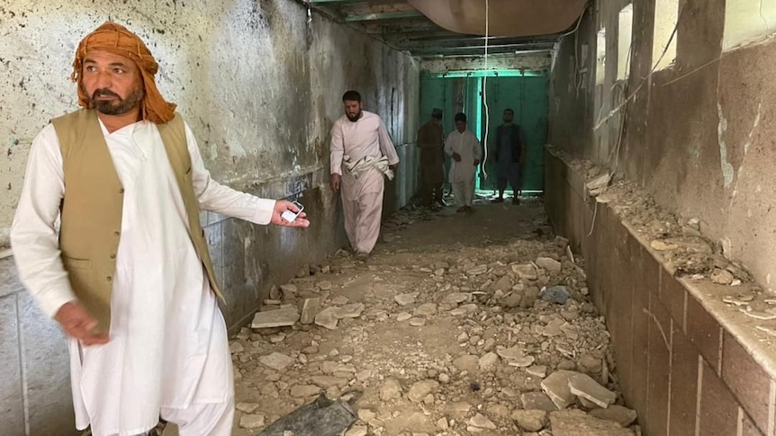 A middle-aged Arabic man in traditional garb points to damage in a stone corridor with men behind.