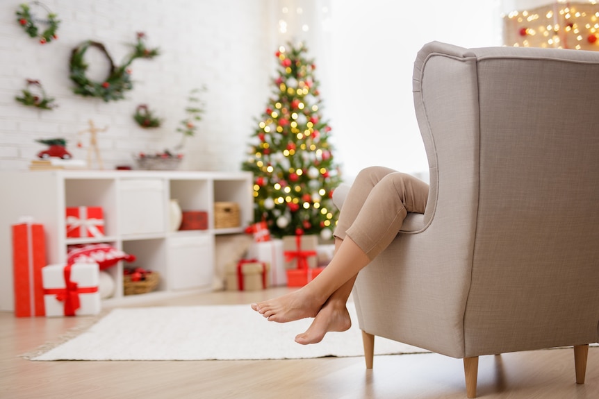 A woman's legs hang over the arm of a grey chair, with a Christmas trees, presents and decorations in the background.