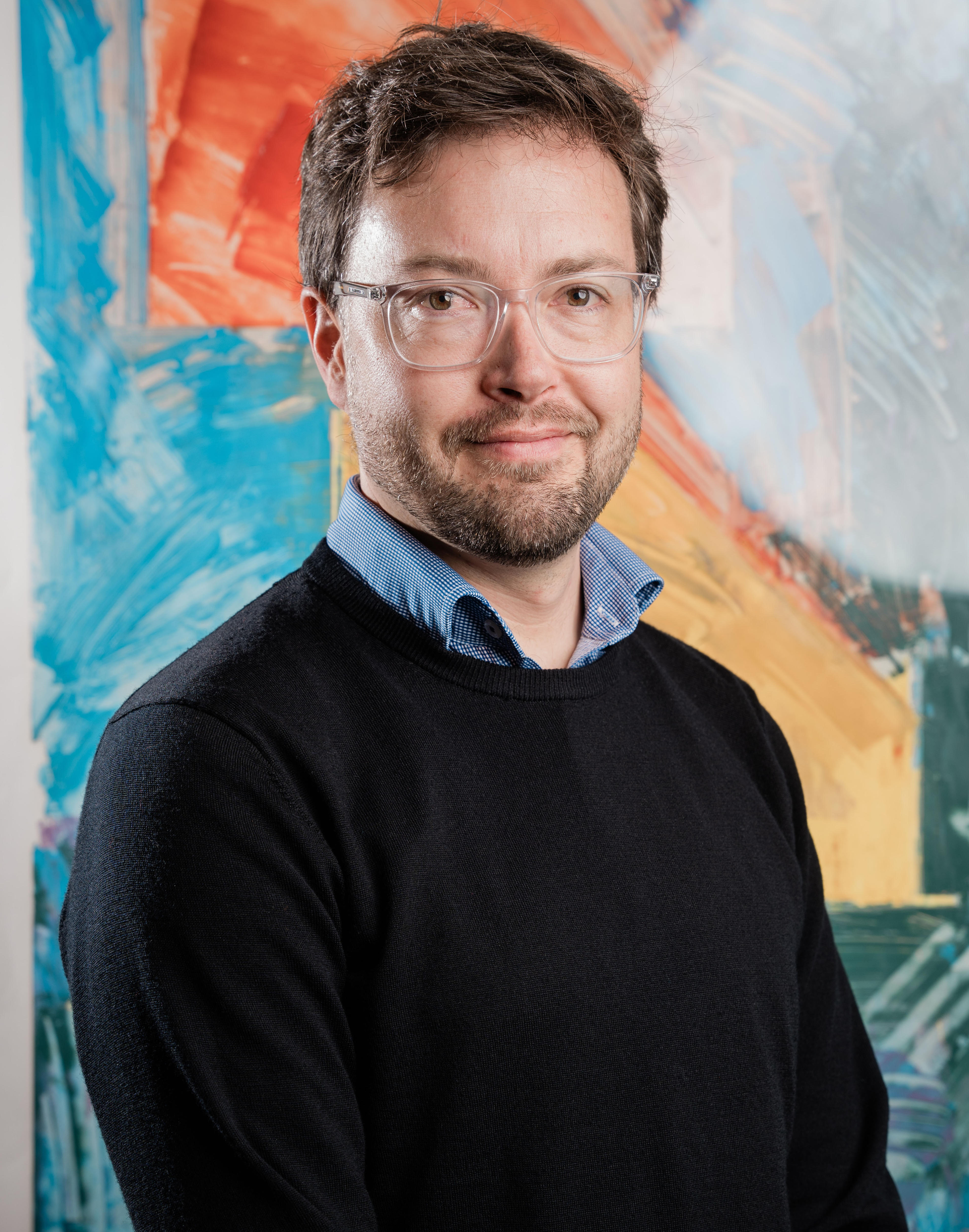 A dark-haired man in aglasses stands in front of a painting.
