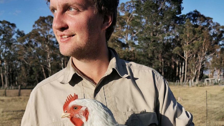 A man is standing near the camera looking away from the camera holding a chicken.