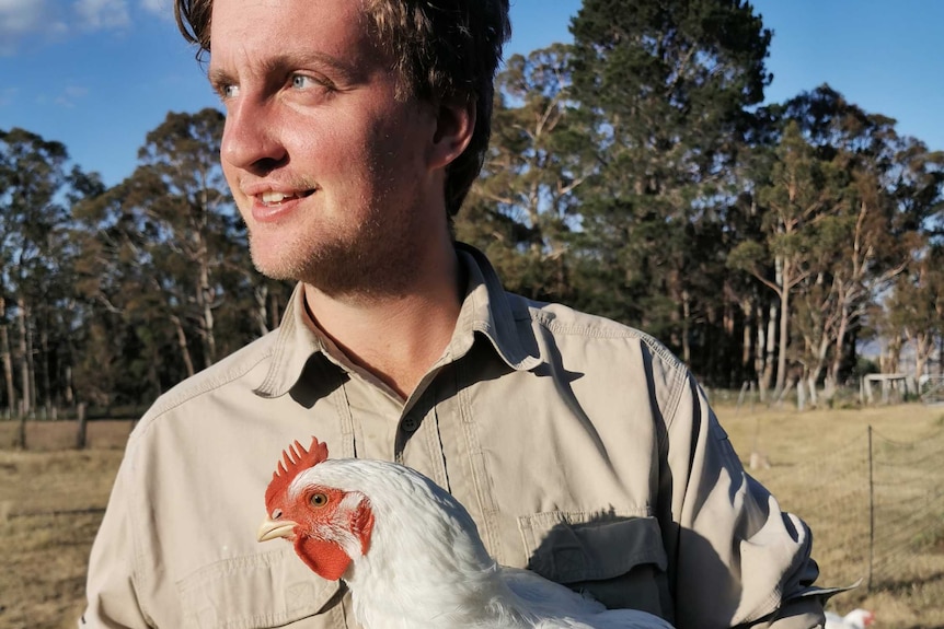 A man is standing near the camera looking away from the camera holding a chicken.