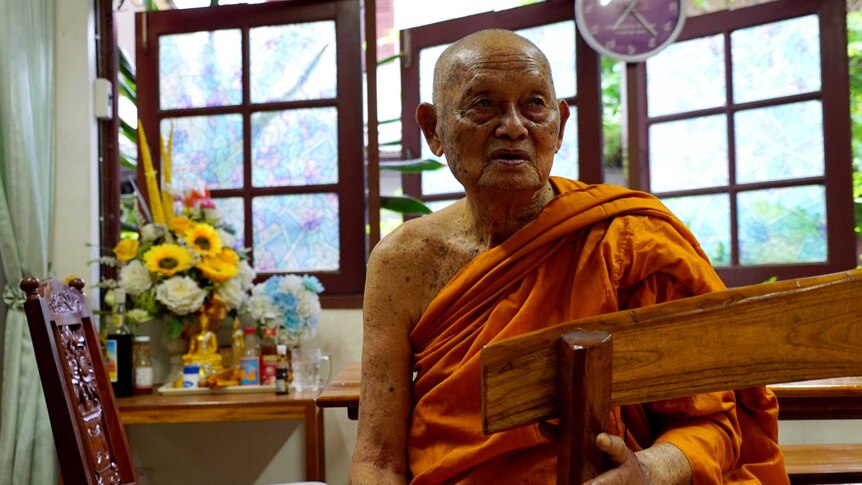 An elderly monk dressed in a saffron robe sits in a room with flowers and a small golden Buddhist statue