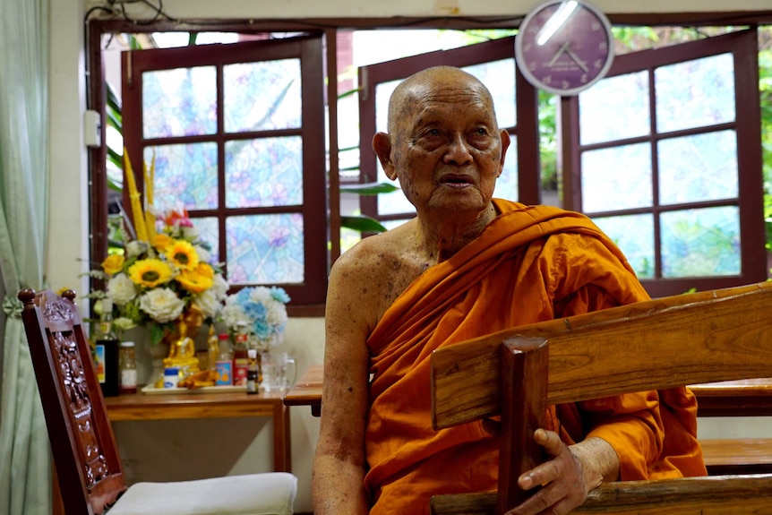 An elderly monk dressed in a saffron robe sits in a room with flowers and a small golden Buddhist statue