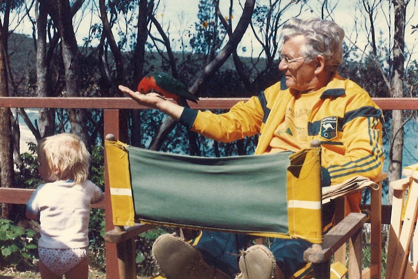 Bob Hawke sits outside with a red and green bird on his hand.