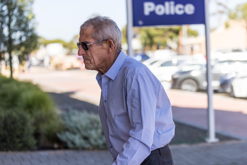 An older man in a business shirt and sunglasses walks toward a court building.