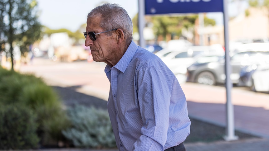 An elderly man in dark sunglasses and a business shirt walks towards an unseen building.