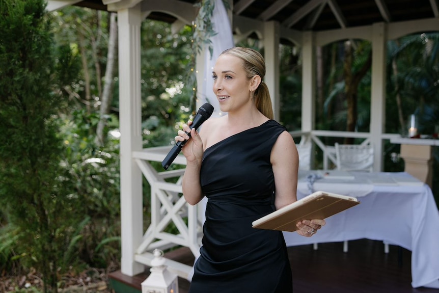 A woman with blonde hair in a black dress speaking into a microphone at a wedding holding an ipad