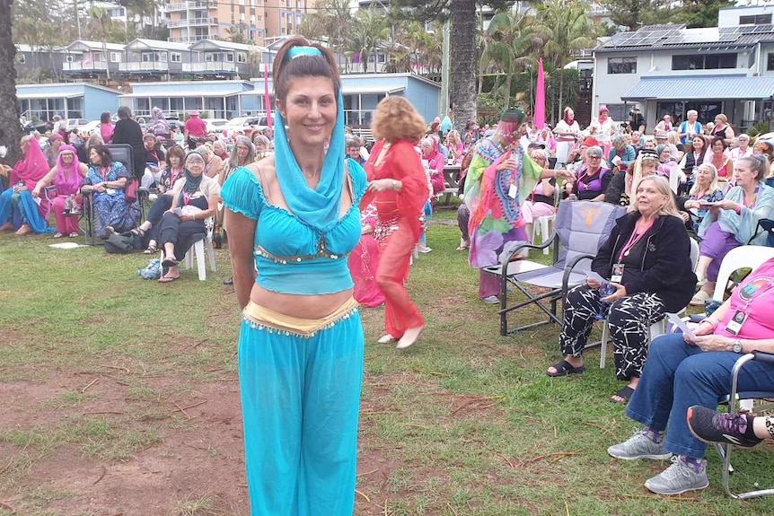 a woman in light blue crop top and harem pants and head scarf in front of a large group of women sitting on chairs