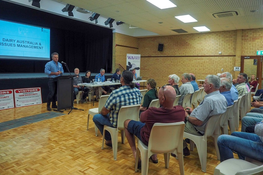 A wide shot showing farmers sitting at a meeting in a hall listening to a man speaking at the front of the room.