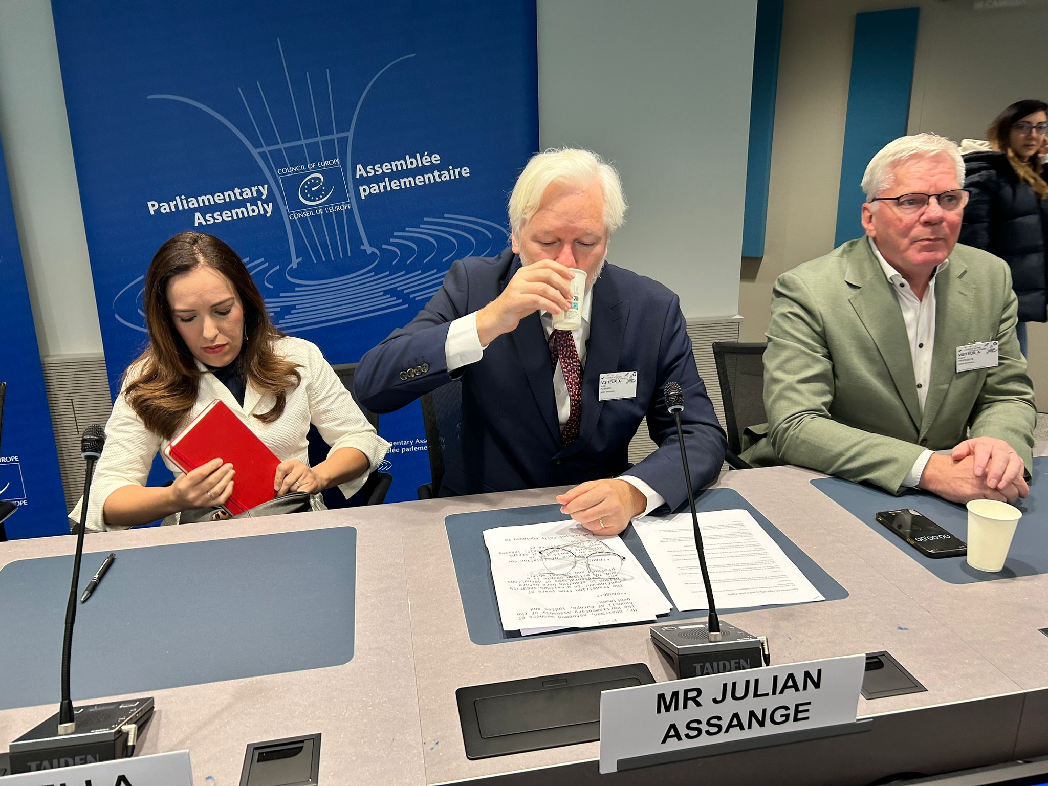 Julian Assange takes a drink of water sitting next to a man and woman. 
