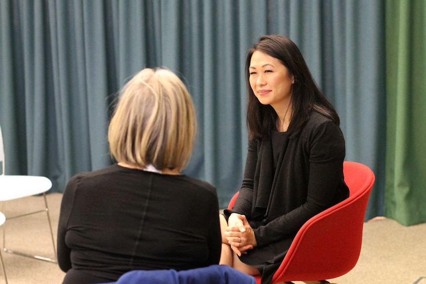 A woman sitting in a red chair talks to a woman whose back is to the camera.