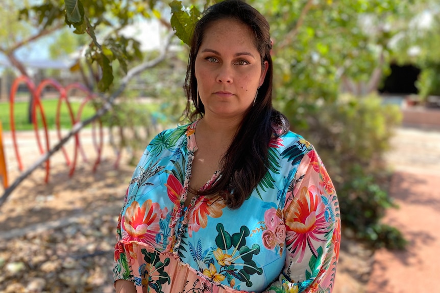 A solemn-looking Indigenous woman with long, dark hair and dressed in a colourful top stands near a shrub.
