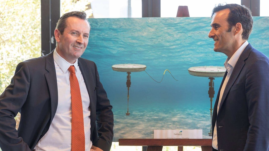 A smiling Mark McGowan stands next to Mike Ottaviano in front of a large poster displaying Carnegie's wave power technology.