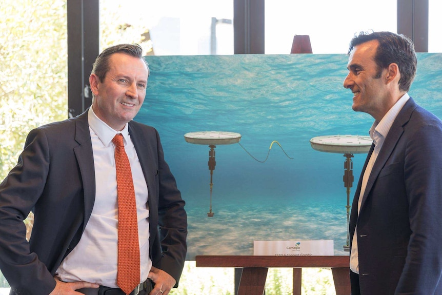 A smiling Mark McGowan stands next to Mike Ottaviano in front of a large poster displaying Carnegie's wave power technology.