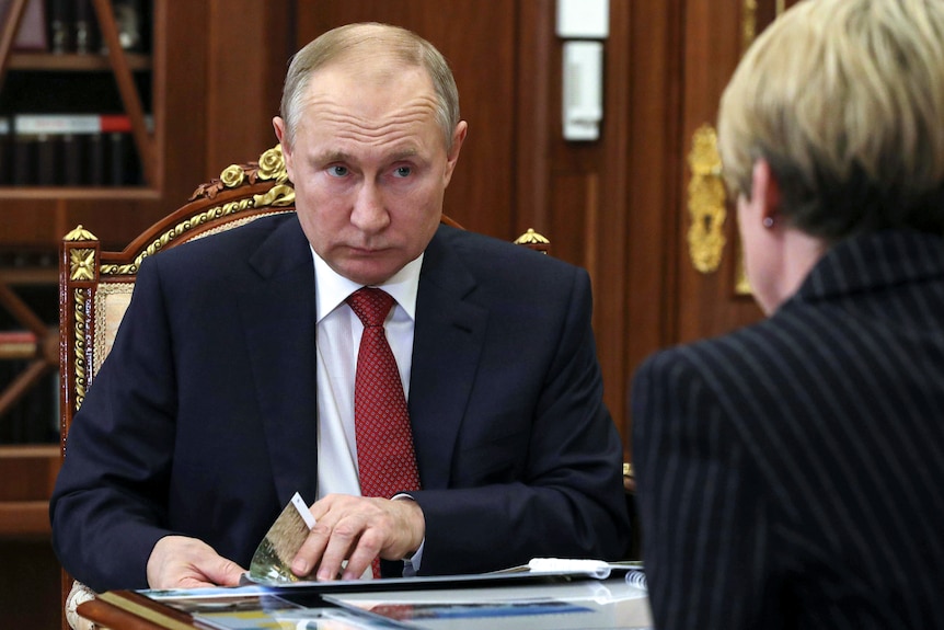 A man sitting on an ornate chair, wearing a suit with a red tie, listening to a woman talk