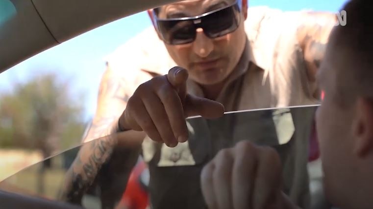 A man wearing sunglasses points through the window of a car while another man watches him from the driver's seat.