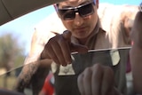 A man wearing sunglasses points through the window of a car while another man watches him from the driver's seat.