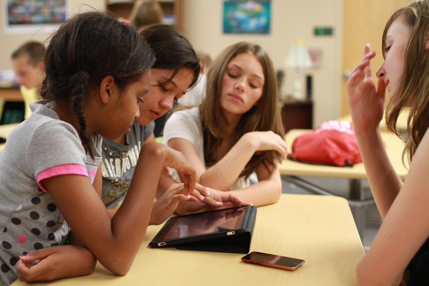Four female students sitting around a desk use a tablet computer in class.
