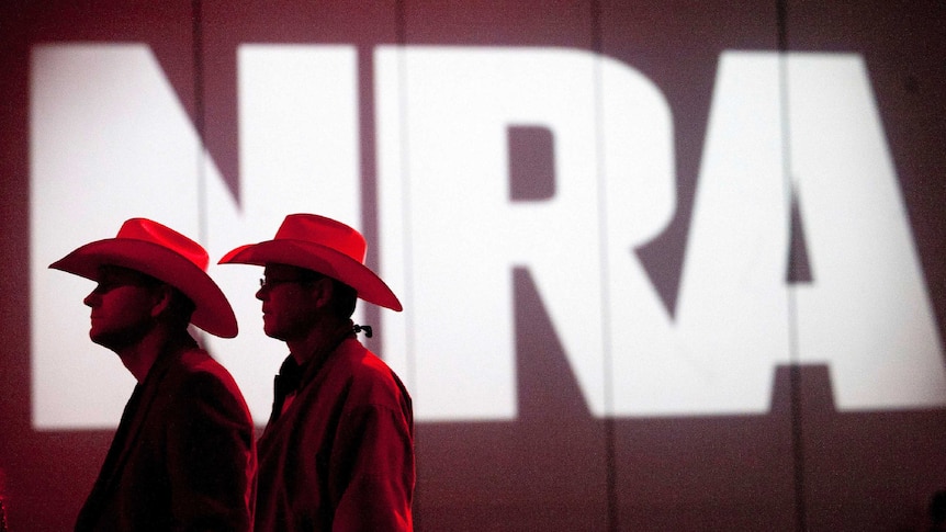 Two men with cowboy hats stand in front of a sign saying NRA.