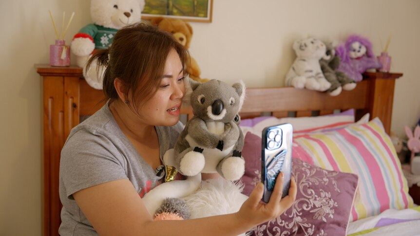 Mother sits on bed holding toy koala up to phone during video call with her children