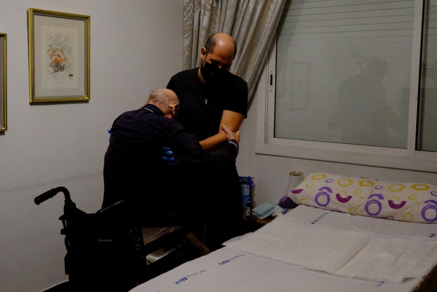 A man wearing a face mask helps another man out of a wheelchair in a bedroom. 