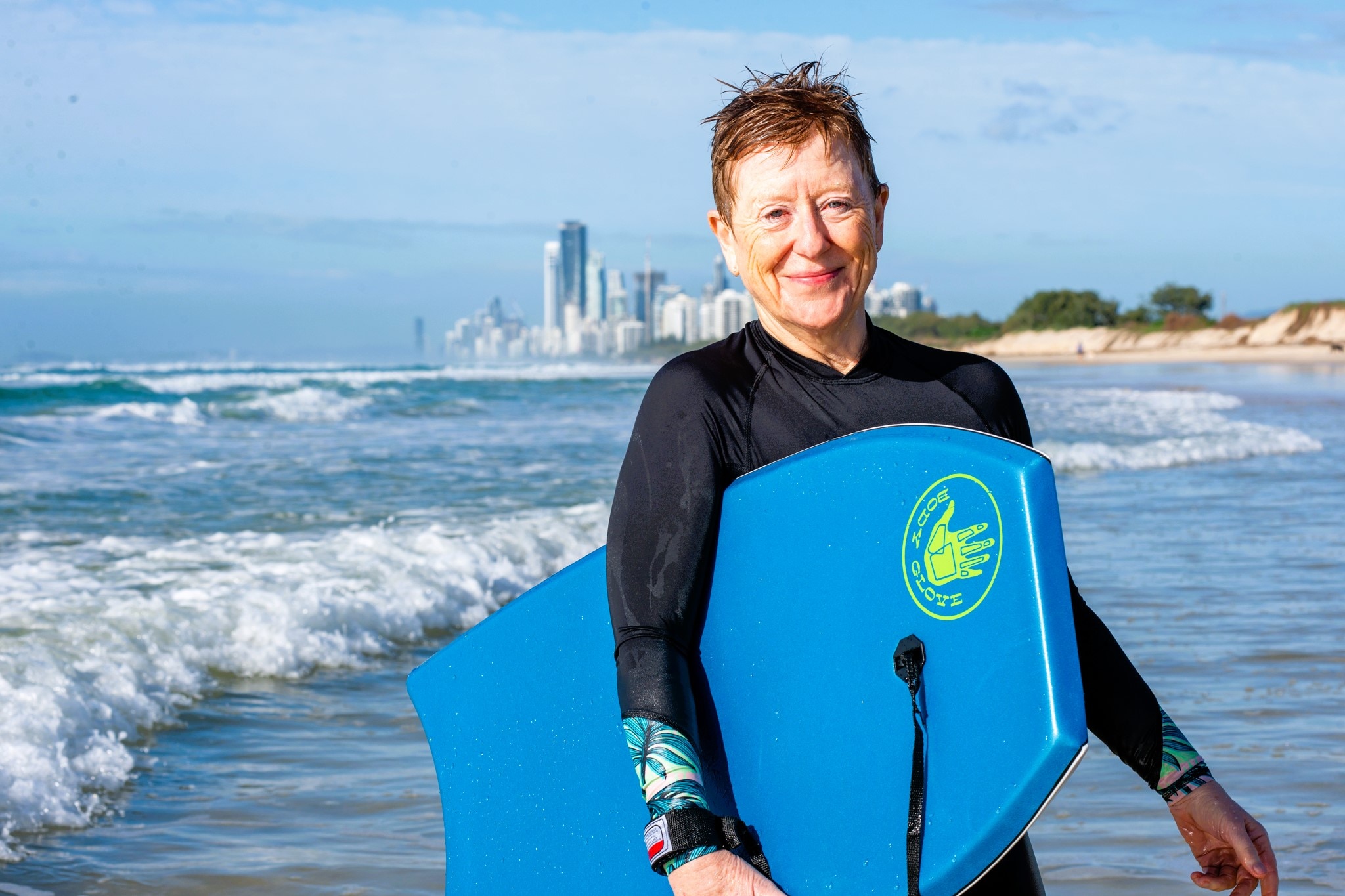 A woman in a black long sleeved surf rashie with a blue boogie board under her arm at the beach.